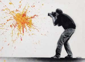 A stencil artwork by Martin Whatson showing a photographer aiming at a vivid orange and yellow paint splatter on a white wall